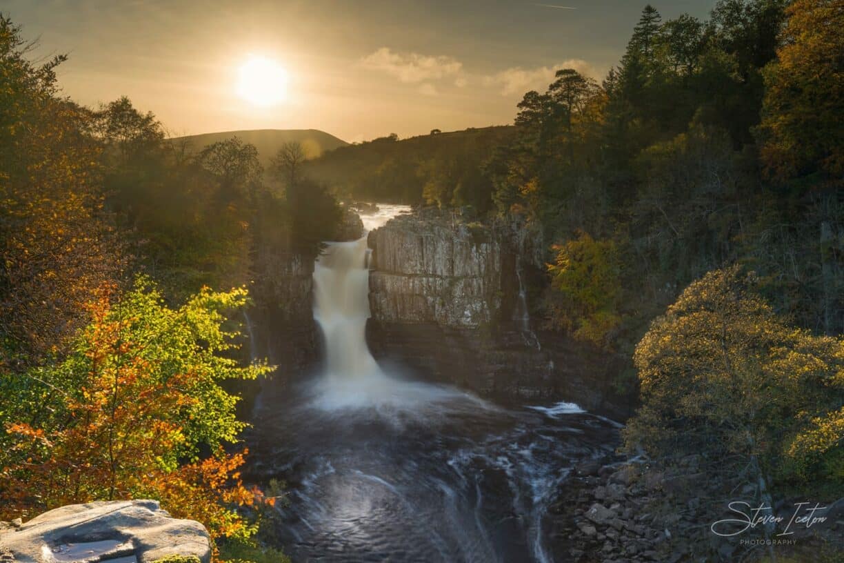 Wide-angle view of High Force Waterfall surrounded by lush green forest under a vibrant summer sunset sky.